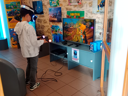 Photo d'une salle de jeu vidéo en bibliothèque. Deux fauteuils font face à deux écrans reliés à des consoles. Deux jeunes garçons de dos jouent chacun à un jeu différent sur les consoles. 