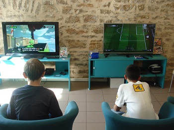 Photo d'une salle de jeu vidéo en bibliothèque. Deux fauteuils font face à deux écrans reliés à des consoles. Deux jeunes garçons de dos jouent chacun à un jeu différent sur les consoles. 